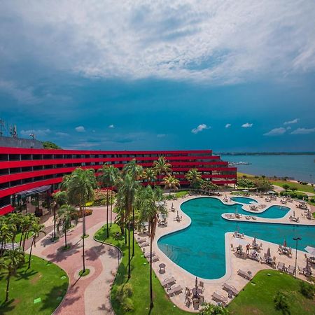 Royal Tulip Brasilia Alvorada Hotel Ngoại thất bức ảnh The photo shows a modern hotel building with a striking red exterior, surrounded by lush greenery. In the foreground, there is a large, creatively shaped swimming pool, with lounge chairs arranged around it. The backdrop features a body of water, pos