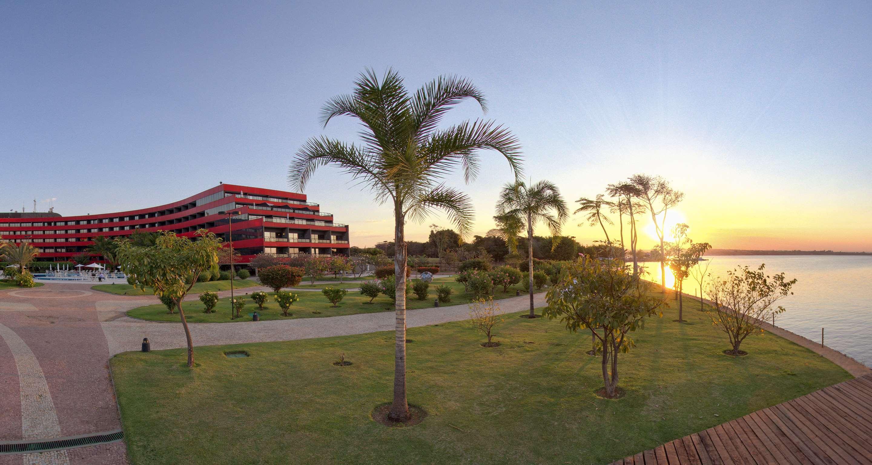 Royal Tulip Brasilia Alvorada Hotel Ngoại thất bức ảnh The photo depicts a scenic view featuring a red multi-story building alongside a serene waterfront. In the foreground, there are several palm trees and lush green lawns, creating a tropical atmosphere. The sun is setting in the background, casting wa