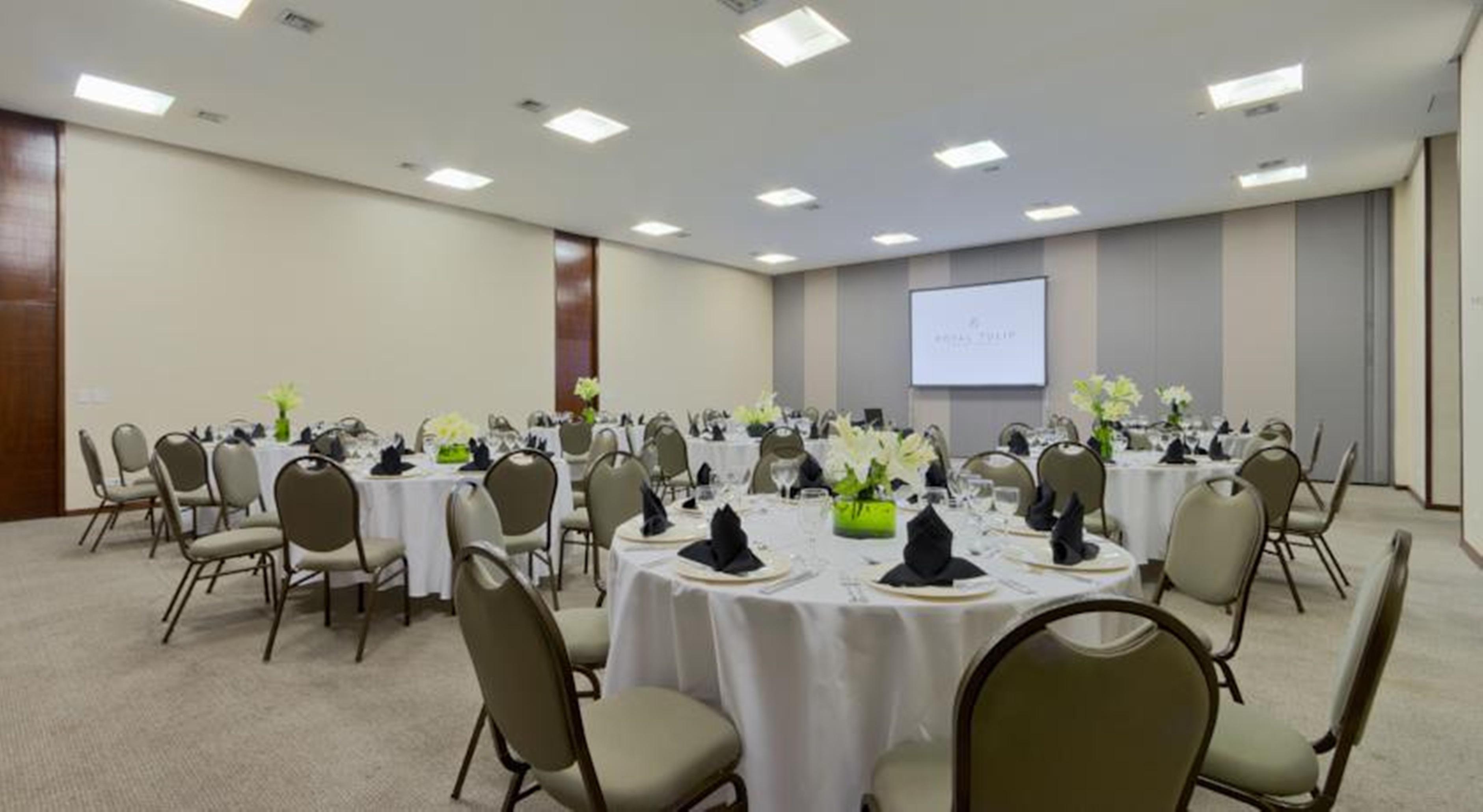 Royal Tulip Brasilia Alvorada Hotel Ngoại thất bức ảnh The photo shows a banquet or conference room set up with multiple round tables covered in white tablecloths. Each table is arranged with black napkins and centerpieces featuring flowers, likely in shades of green and white. The room has beige walls a