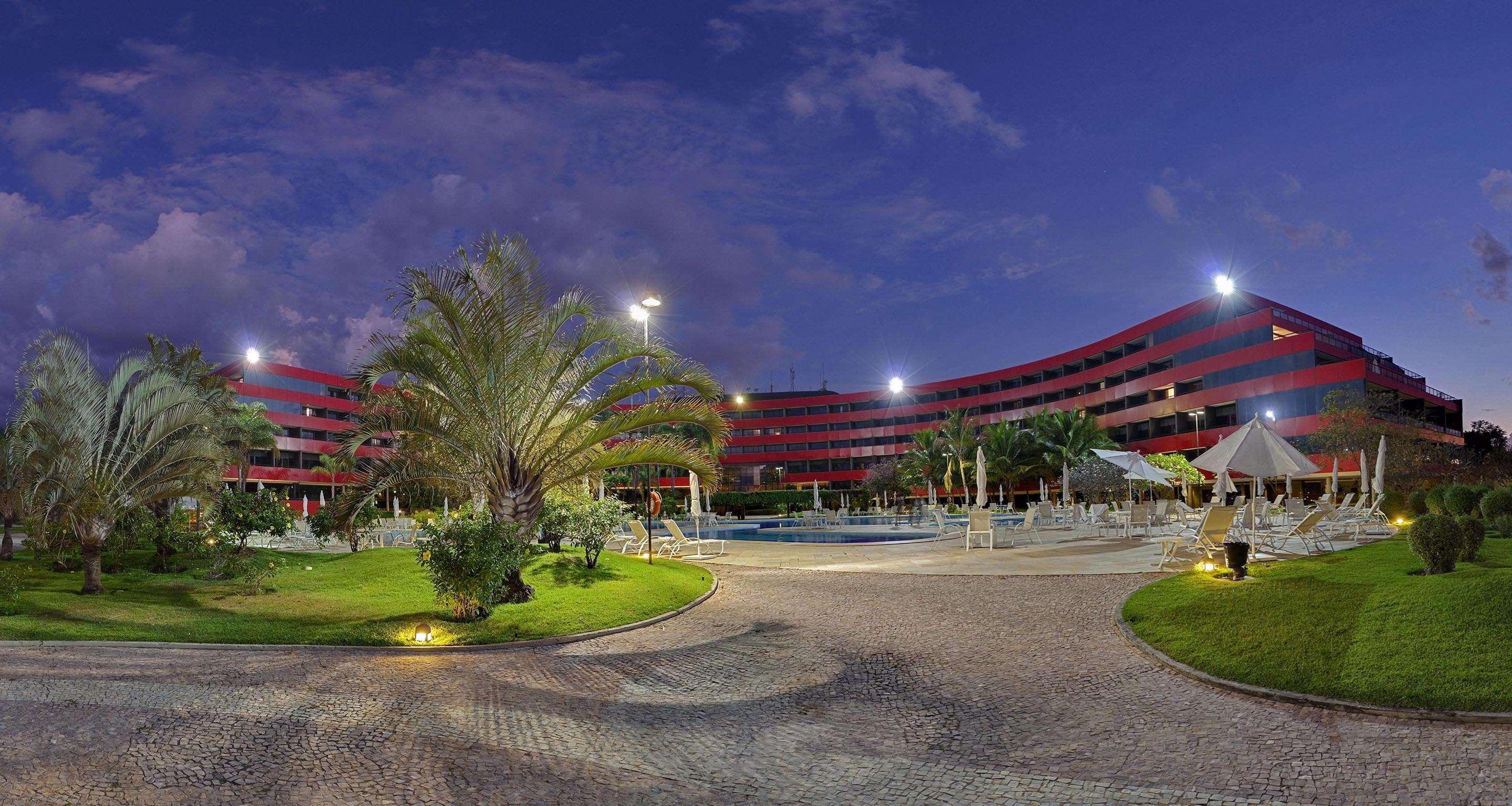 Royal Tulip Brasilia Alvorada Hotel Ngoại thất bức ảnh The photo shows a panoramic view of a hotel or resort complex, characterized by its modern architectural design with smooth, curved lines and a vibrant red color. It’s set in a landscaped area featuring palm trees and green grass. The surroundings in