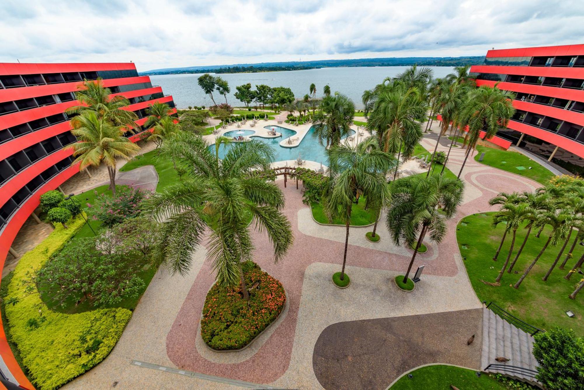 Royal Tulip Brasilia Alvorada Hotel Ngoại thất bức ảnh The photo shows a scenic view of a courtyard area in a residential complex. There are multiple red buildings surrounding a pool area, which is surrounded by palm trees and landscaped gardens. The pool features a curvy design, and there are pathways m