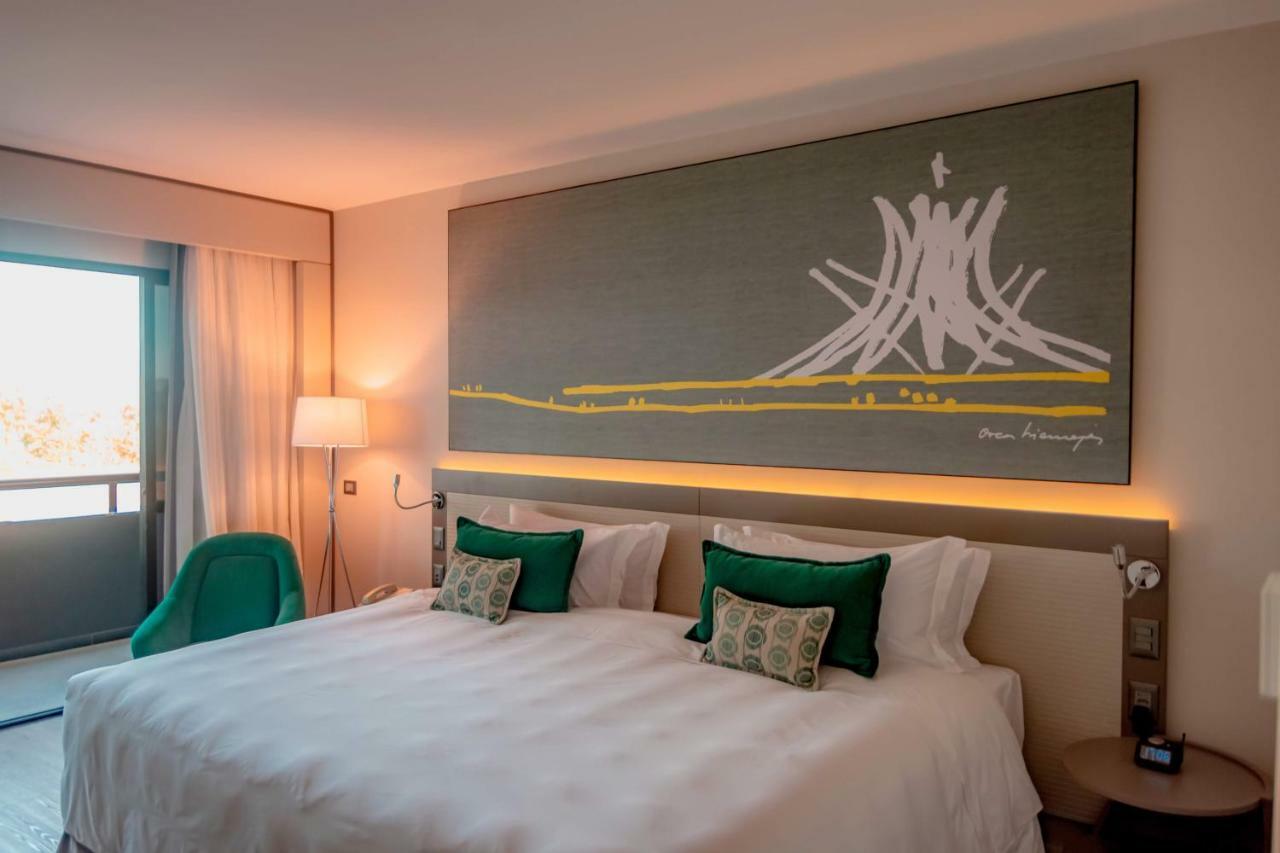 Royal Tulip Brasilia Alvorada Hotel Ngoại thất bức ảnh The photo shows a modern hotel room featuring a large bed with white bedding and two green accent pillows. Above the bed, there is a large piece of artwork depicting a stylized mountain or volcanic shape with yellow accents. The room has a contempora